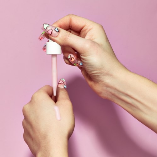Hände mit bunten Herz-Nageldesign halten eine rosa Lippenstiftpinsel vor rosa Hintergrund.