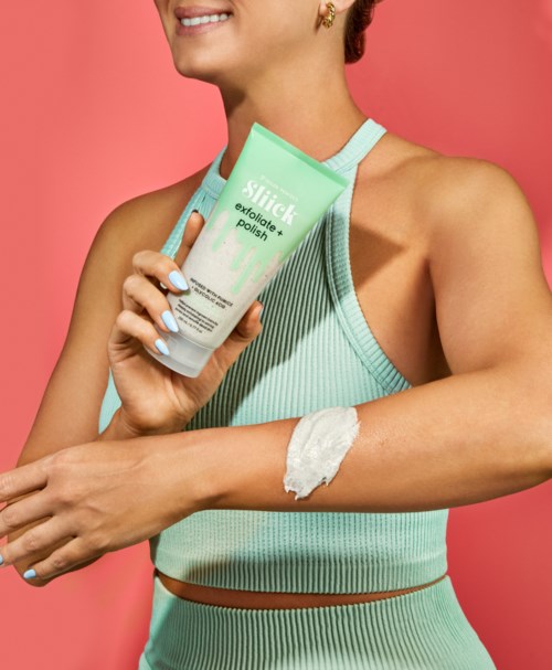 Eine Frau in Sportkleidung trägt Creme auf den Arm auf, hält eine Tube mit Peeling, lächelt.