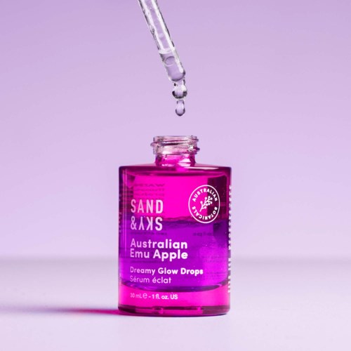 Eine pinke und lila Flasche mit 'Sand & Sky'-Aufdruck, mit Pipette und Tropfen darüber, vor helllila Hintergrund.