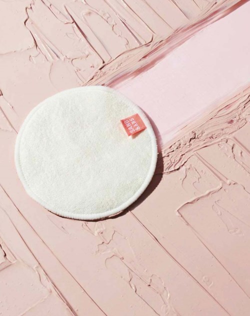 Ein weißer runder Make-up-Schwamm mit einem kleinen roten Tag auf einer strukturierten rosa Platte.