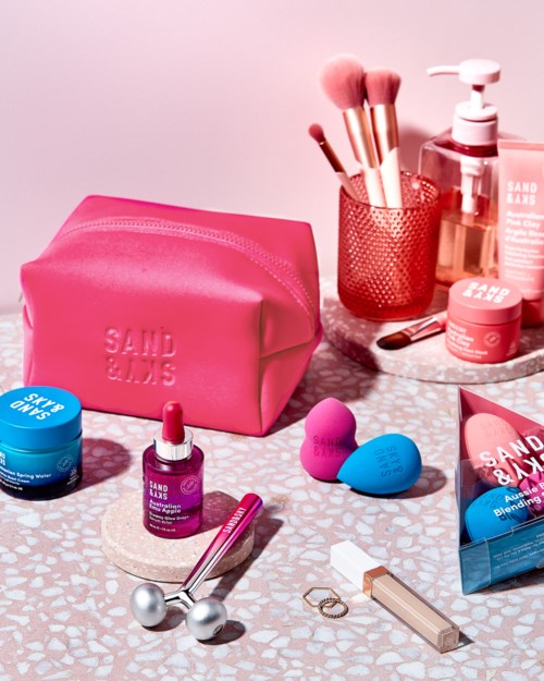 Schönheitspflegeprodukte und Werkzeuge, darunter Make-up-Pinsel, Schwämme, Lippenstift, Hautpflege und eine pinke Tasche, auf einer gemusterten Oberfläche angeordnet.