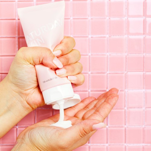 Een persoon die witte lotion uit een tube op hun handpalm drukt tegen een roze betegelde achtergrond.