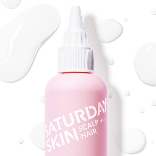 Een roze fles met witte dop gemerkt "Saturday Skin Scalp + Hair". Het ligt op een witte achtergrond met enkele druppels.