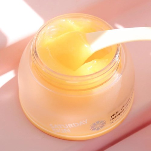 Een gele crème of gel in een pot met een witte spatel die eruit steekt. De pot heeft tekst en een kleine citrusafbeelding.