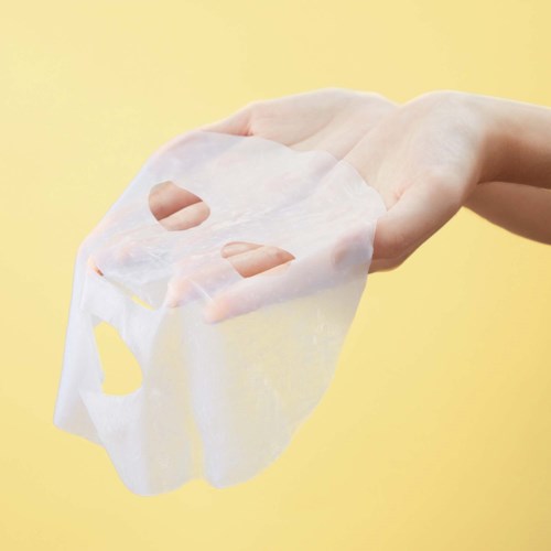 Een hand houdt een wit gezichtsmasker tegen een gele achtergrond.