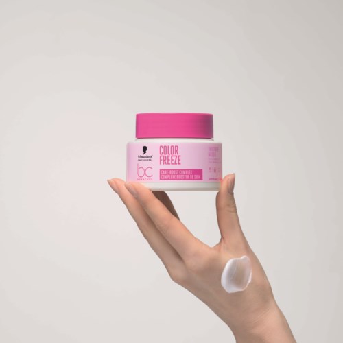 En hånd holder en rosa krukke merket color freeze, med krem eller lotion på hånden, mot en enkel bakgrunn.