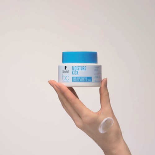 Käsi pidittää sinistä ja valkoista purkkia nimeltä Moisture Kick, jossa on pieni määrä voidetta sormella.