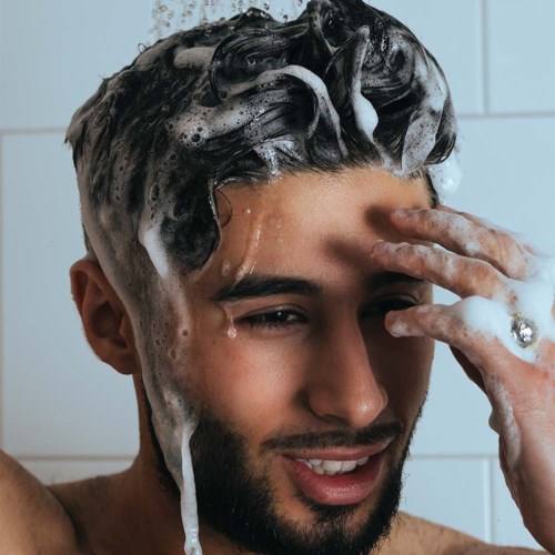 Ein Mann wäscht sich die Haare mit Seife, Schaum und Blasen, lächelnd in der Dusche.