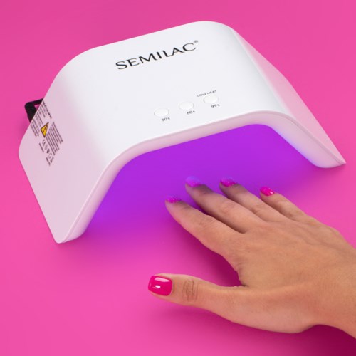 Eine Hand unter einer weißen UV-Nagellampe mit violettem Licht auf einer pinken Oberfläche. Die Lampe hat Tasten mit den Aufschriften 10, 30 und 60.