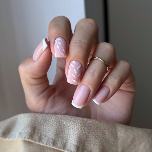 A hand shows nails with a pink base, white tips, and delicate white leaf patterns. The hand wears a ring and is near a beige fabric.