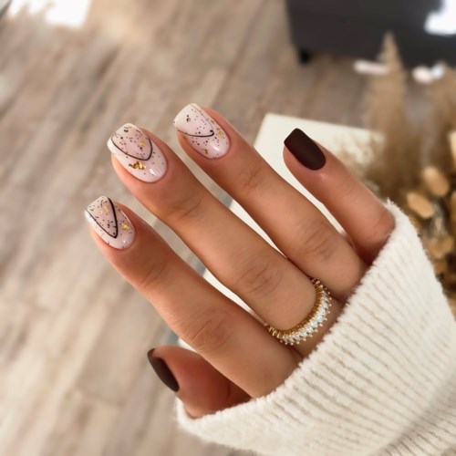 Nägel sind mit weißem Lack, schwarzen Linien, Goldpunkten und Glitzer verziert. Hand trägt einen Ring und einen cremefarbenen Pullover.