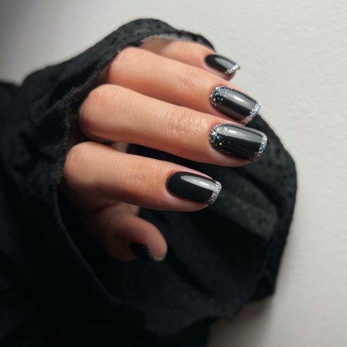 Fingernägel mit schwarzem Nagellack und silbernem Glitter, teilweise bedeckt von dunklem Stoff.
