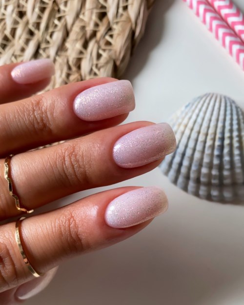 Nail polish with a subtle glittery finish on natural-looking, oval-shaped nails. Decorations include a shell and a straw.