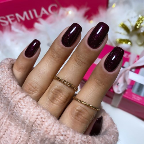 Nahaufnahme einer Hand mit dunkelroten, glänzenden, glitzernden Nägeln, zwei Goldringen, im pinken festlichen Hintergrund.
