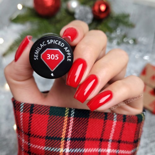 Eine Hand mit roten Nägeln hält einen schwarzen Lippenstift mit der Beschriftung "Semillac Spiced Apple 305" vor einem Weihnachtsbaum.