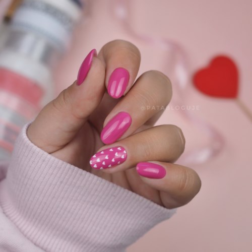Eine Hand mit pinkfarbenen und gemusterten Nägeln vor einem Herz und Nagellackflaschen auf rosa Hintergrund.