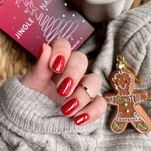 Hand mit rotem Nagellack, grauer Pullover, Lebkuchen-Ornament und Weihnachtskarte im Hintergrund.