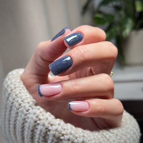 Nail polish with a sparkly blue design on some nails, and light pink with blue tips on others. Hand near a cozy sweater.