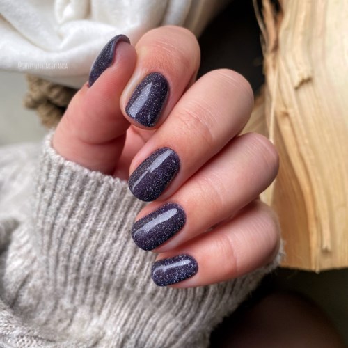 Nails painted with dark, glittery purple nail polish. Hand is partially covered by a grey sweater. Background has wood.