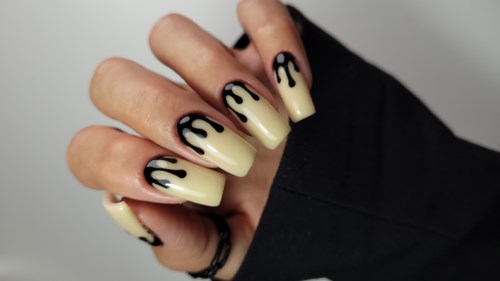 Nageldesign mit schwarzer Tropfenmuster auf gelben Nägeln. Hand ist teilweise mit schwarzem Ärmel oder Handschuh bedeckt.