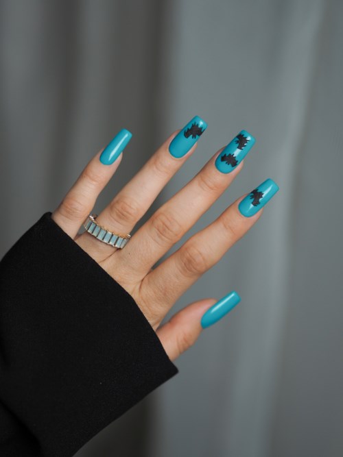 Eine Hand mit blauer Maniküre und schwarzen Fledermaus-Designs, trägt einen Silberring vor grauem Hintergrund.