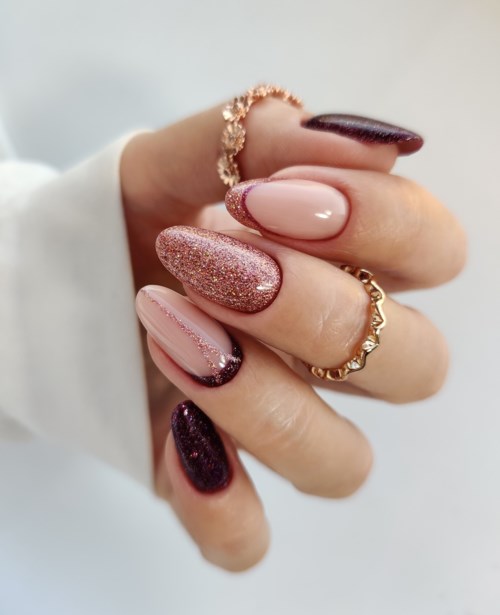 A hand with decorated nails and gold rings, featuring pink, glitter, and dark purple nail polish.
