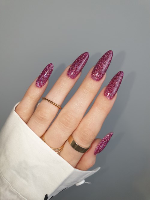 Hand mit langen, glitzernden pinken Nägeln, Ringen und weißem Ärmel vor einfachem Hintergrund.