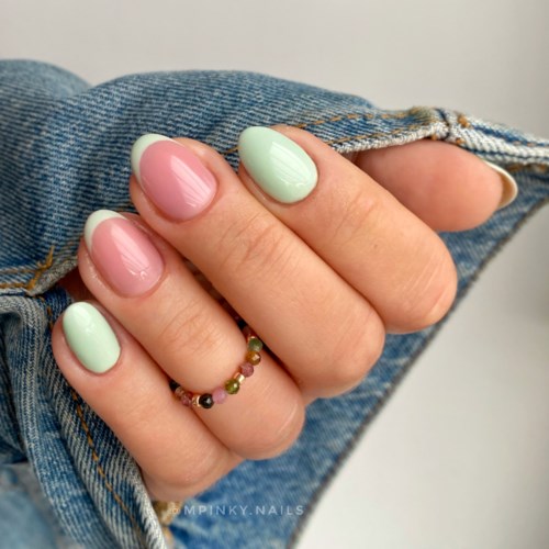 Nägel in Pink und Grün lackiert, mit einem goldenen Ring mit kleinen bunten Steinen, gehalten vor Jeansstoff.