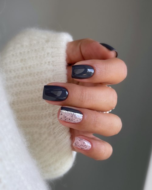 Hand mit verzierten Nägeln, einige dunkelgrau und einige mit rosa Glitzer, trägt einen weißen Pullover.
