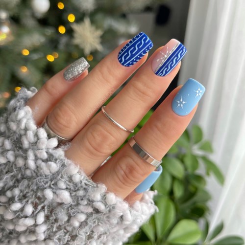 Nägel sind mit blauen, silbernen und weißen Mustern und Schneeflocken verziert, gehalten von einer Hand nahe eines Weihnachtsbaums.
