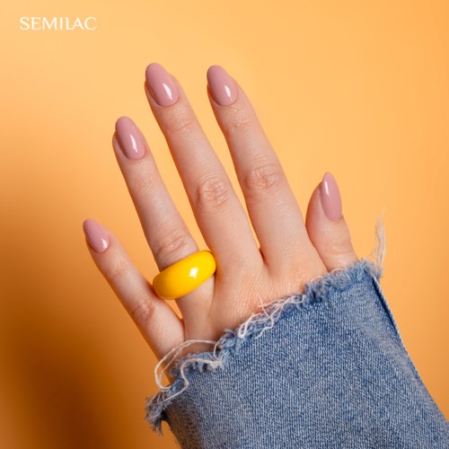 Eine Hand mit rosa Nägeln und einem gelben Ring, in einer Jeansärmeln, vor einem pfirsichfarbenen Hintergrund.