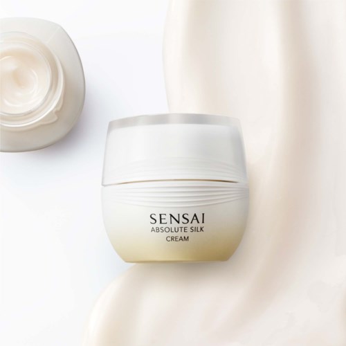 Ein weißer Behälter mit der Aufschrift Sensai Absolute Silk Cream, umgeben von Creme auf einer weißen Oberfläche.