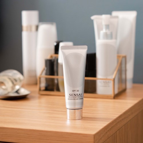 Ein weißer Tuben von Sensai SPF 30 Sonnenschutzcreme auf einer Holzoberfläche mit verschiedenen Hautpflegeflaschen im Hintergrund.