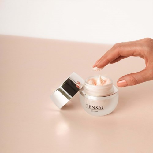 Een hand opent een witte pot met roze crème erin, met label 'SenSae'.