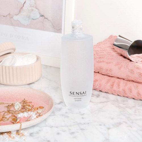 Een duidelijke fles Sensai-lotion op een marmeren oppervlak, met roze handdoeken, sieraden en huidverzorgingsproducten eromheen.