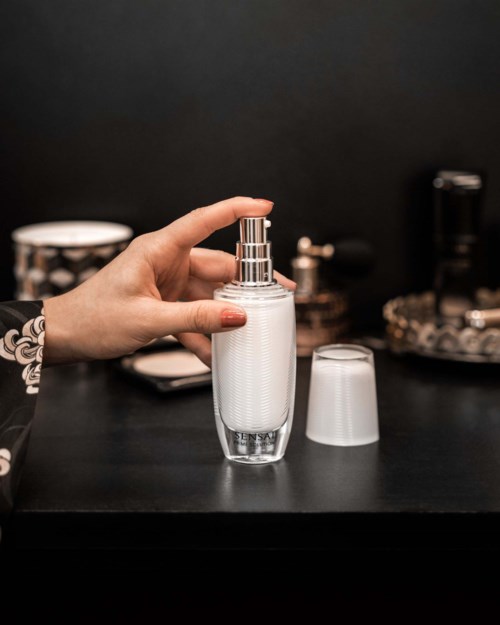Een hand houdt een doorzichtige sprayfles met een zilveren pomp, naast een plastic deksel op een donkere ondergrond.