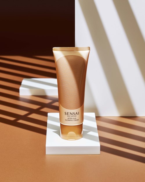A tube of Sensai skincare cream on a white stand, with shadows and sunlight creating patterns around it.