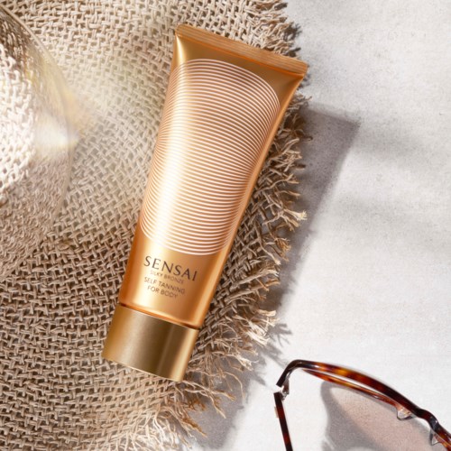 Een goudkleurige tube Sensai SPF 50 zonnebrandcrème op een gestructureerd doek, naast een zonnebril.