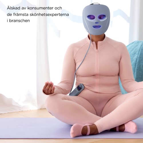 A person sitting in a meditative pose wearing a purple mask, pink clothes, and holding a device, indoors.