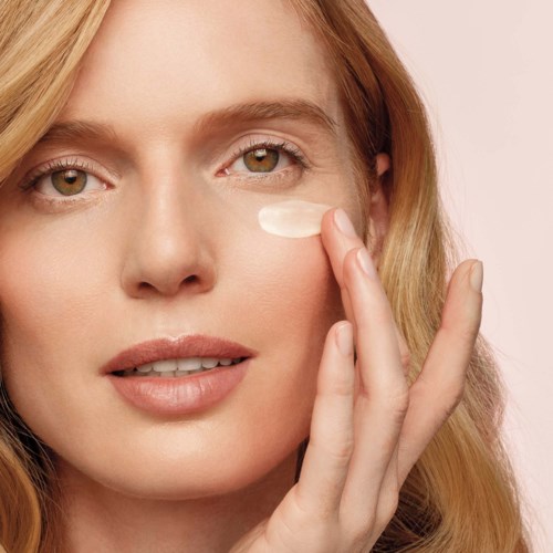 Vrouw brengt crème aan onder haar oog, met lichte make-up en lang blond haar, ziet er kalm en natuurlijk uit.
