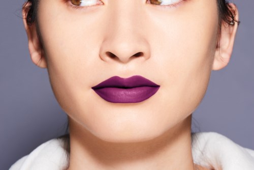 Närbild av en persons ansikte med lila läppstift och minimal makeup, grå bakgrund.