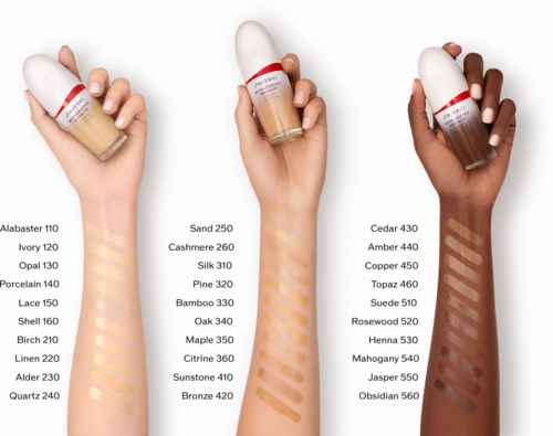 Three hands hold foundation bottles with different skin tones and shades, displaying swatches of various makeup colors.
