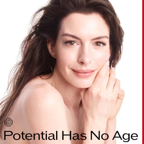 A woman with smooth skin and dark brown hair gently touching her face, smiling softly against a plain background.