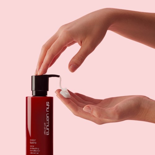 Een hand druppelt shampoo uit een rode fles met zwarte dop op een roze achtergrond.