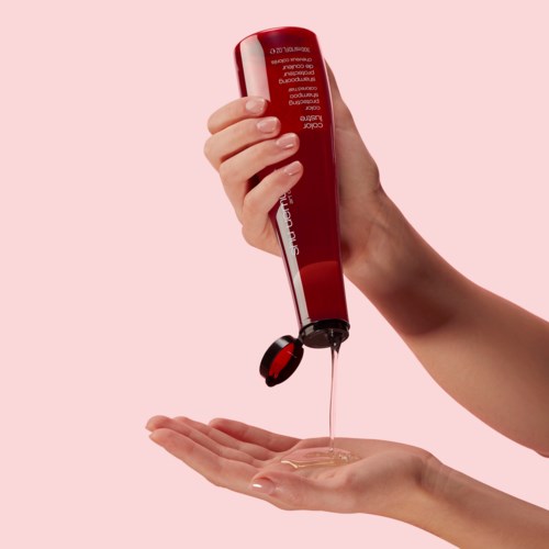 Eine Hand hält eine rote Flasche, aus der eine klare Flüssigkeit auf eine andere Hand gegossen wird, vor rosa Hintergrund.