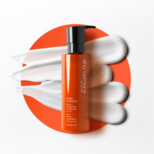 Eine Flasche Shu Uemura Urban Moisture Conditioner mit weißen Creme umgeben und einem orangefarbenen Hintergrund.