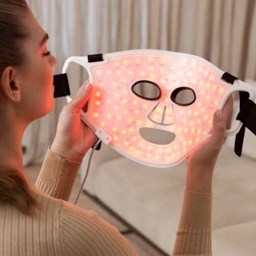 Eine Frau hält eine Gesichtsmaske mit LED-Lichtern, um die Haut zu verbessern, in einem gemütlichen Innenraum.