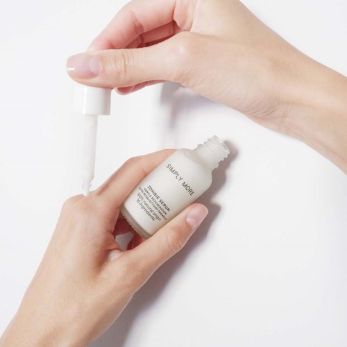 A hand applies white nail care serum from a dropper bottle onto nails.