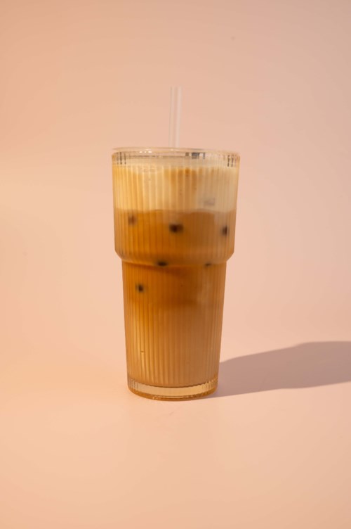 Glass med kald kaffe med melk og isbiter, sugerør, på lys bakgrunn.