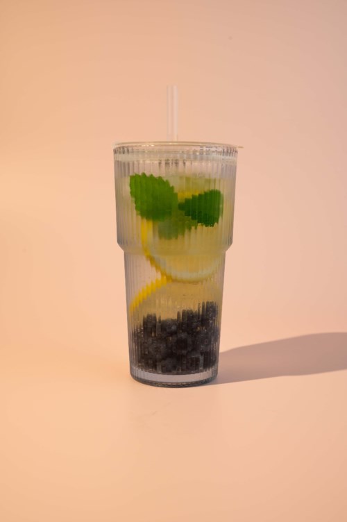 Et langt glass med infusjon av vann med sitronskiver, mynteblader og blåbær med et sugerør mot en lys bakgrunn.